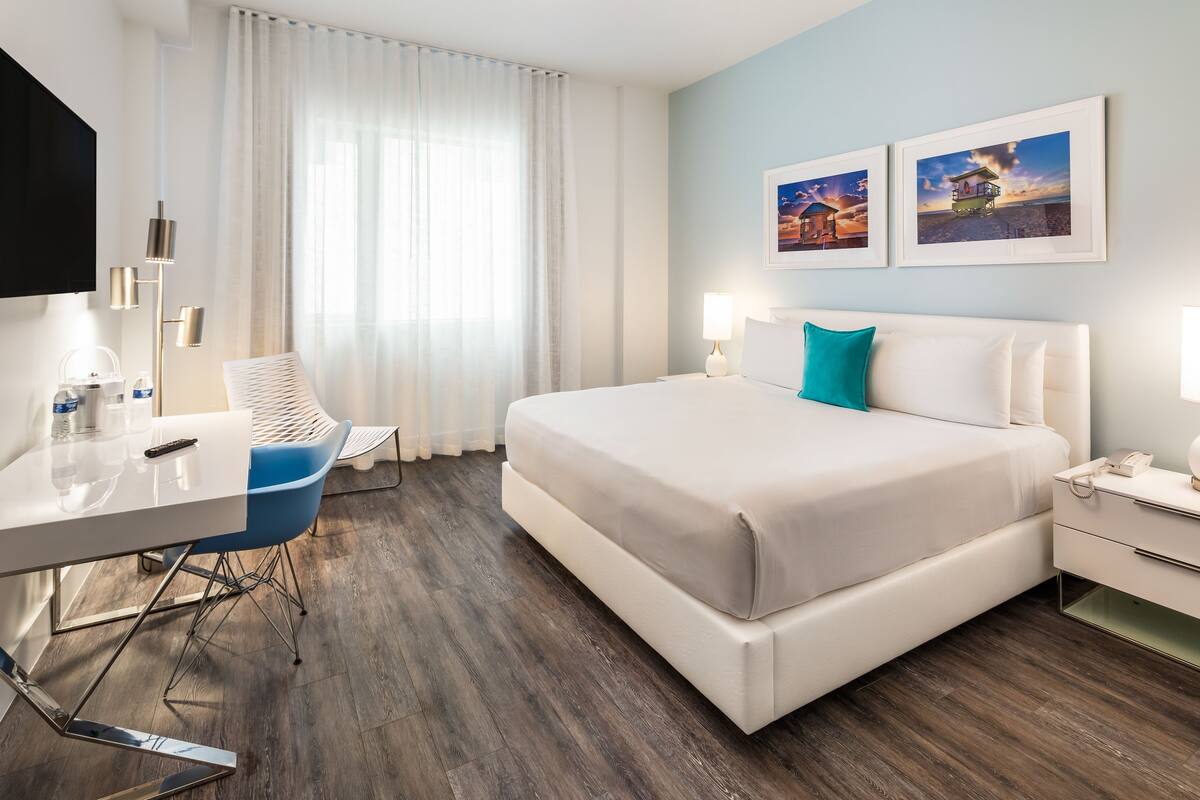A modern hotel room with a large bed, a desk with a chair, a bedside table, and a TV mounted on the wall.