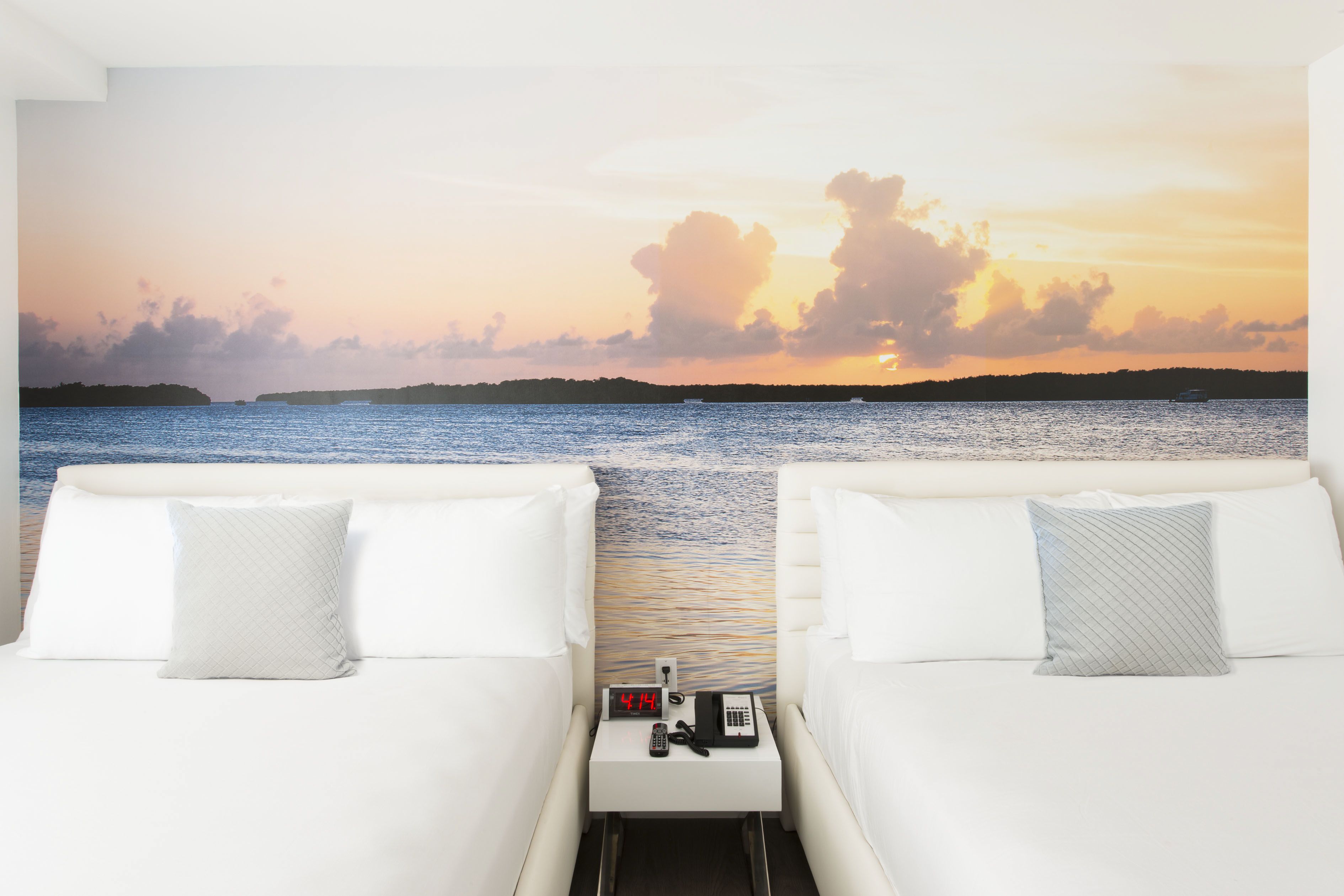 A hotel room with two beds, a nightstand with a clock and phone, and a mural of a sunset over water.