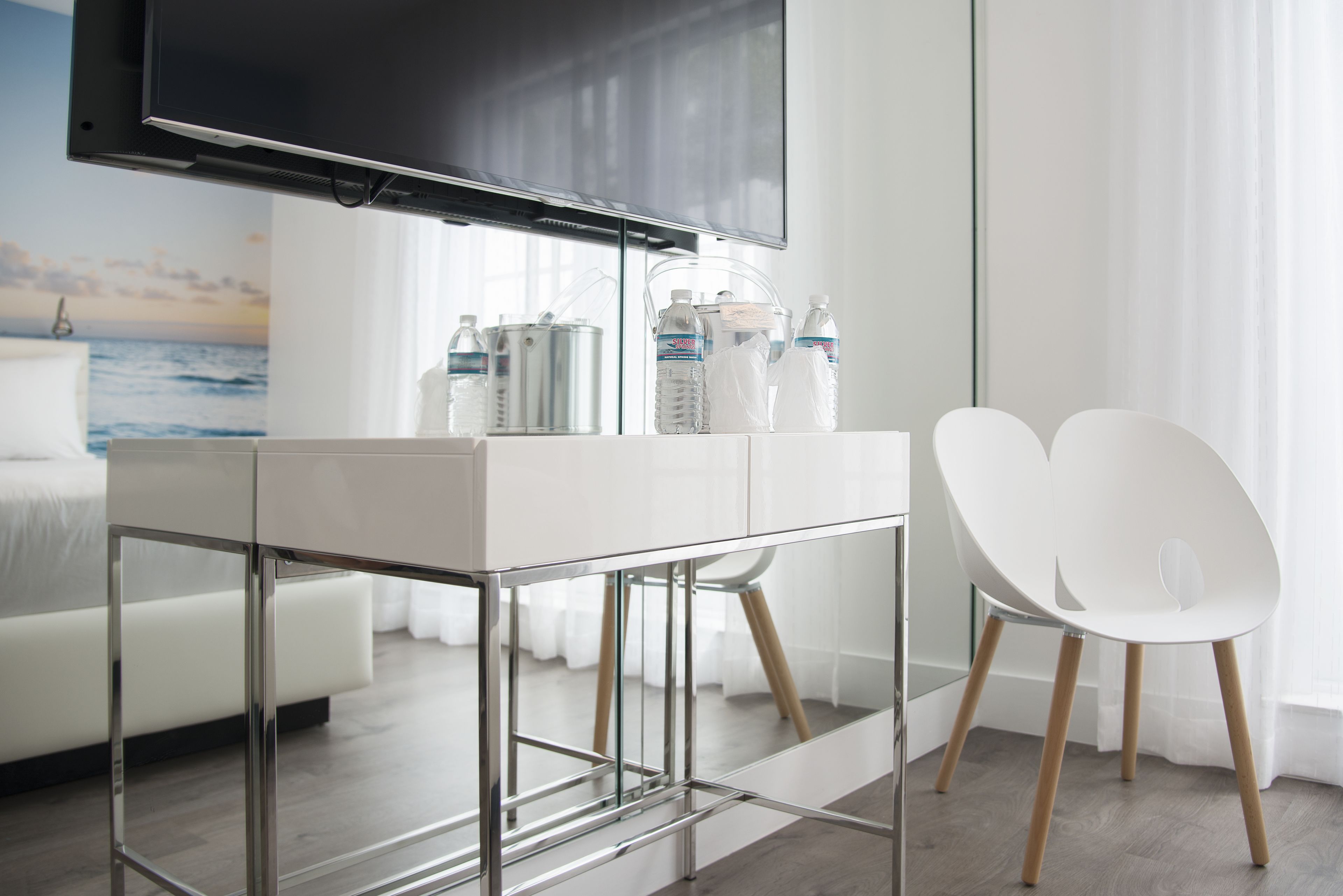 Une chambre d’hôtel moderne dotée d’un grand téléviseur à écran plat fixé au mur, d’un bureau blanc au plateau en verre, de deux chaises blanches et d’un lit avec une tête de lit blanche.