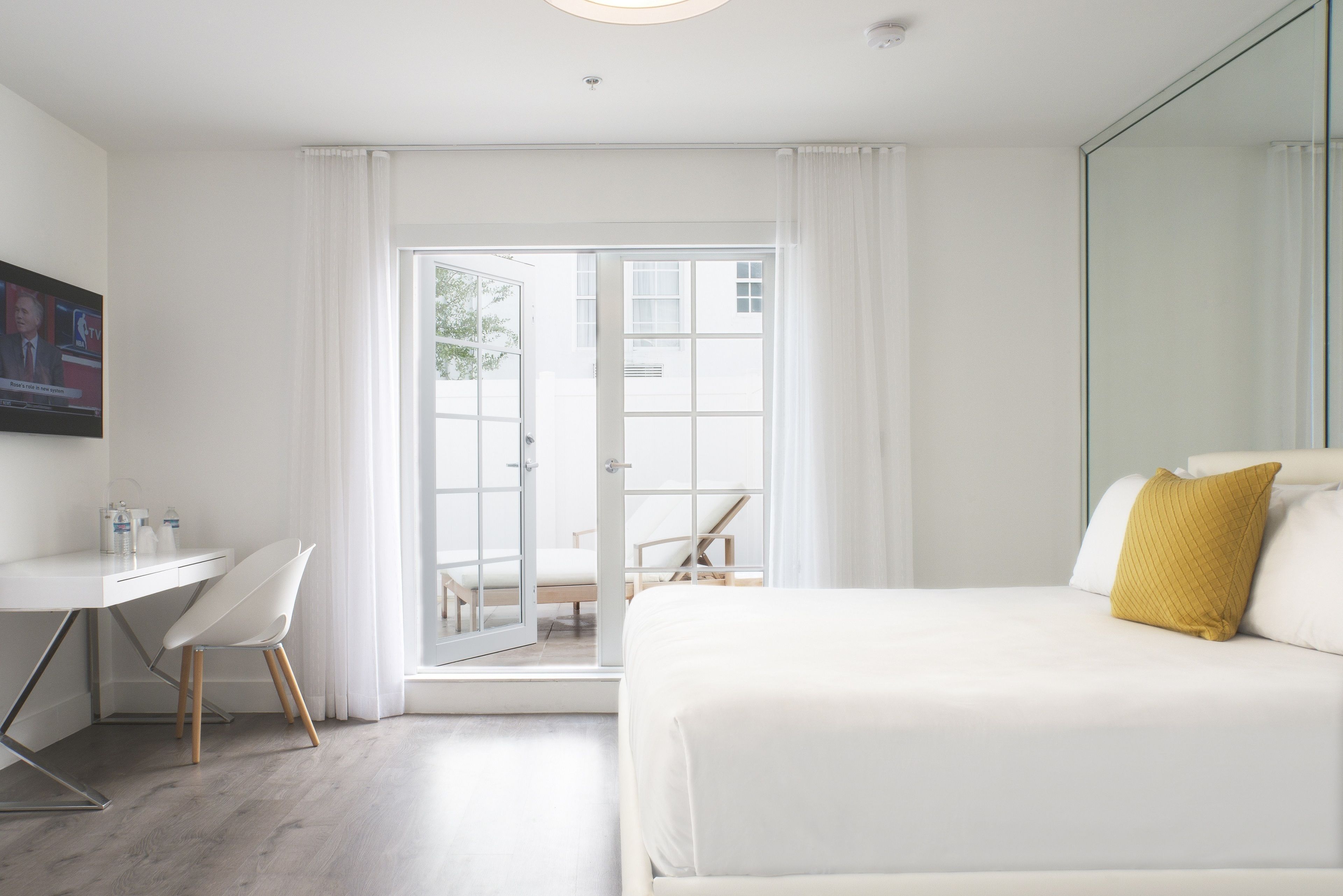 A modern hotel room with a white bed, a desk with a chair, and a large window with curtains.
