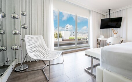 A modern hotel room with a large bed, a white chair, a desk, a flat-screen TV, and a balcony with a view of the city.
