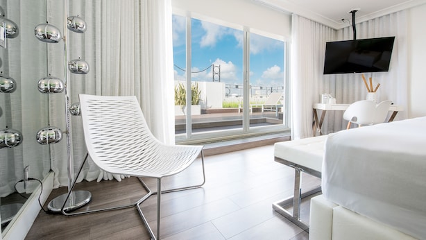 Une chambre d’hôtel moderne dotée d’un grand lit, d’une chaise blanche, d’un bureau, d’une télévision à écran plat et d’un balcon offrant une vue sur la ville.