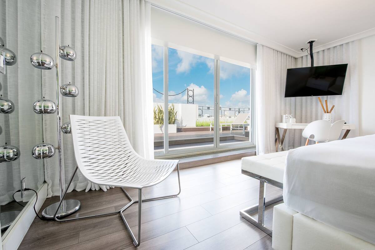 A modern hotel room with a large bed, a white chair, a desk, a flat-screen TV, and a balcony with a view of the city.