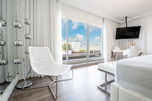 A modern hotel room with a large bed, a white chair, a desk, a flat-screen TV, and a balcony with a view of the city.