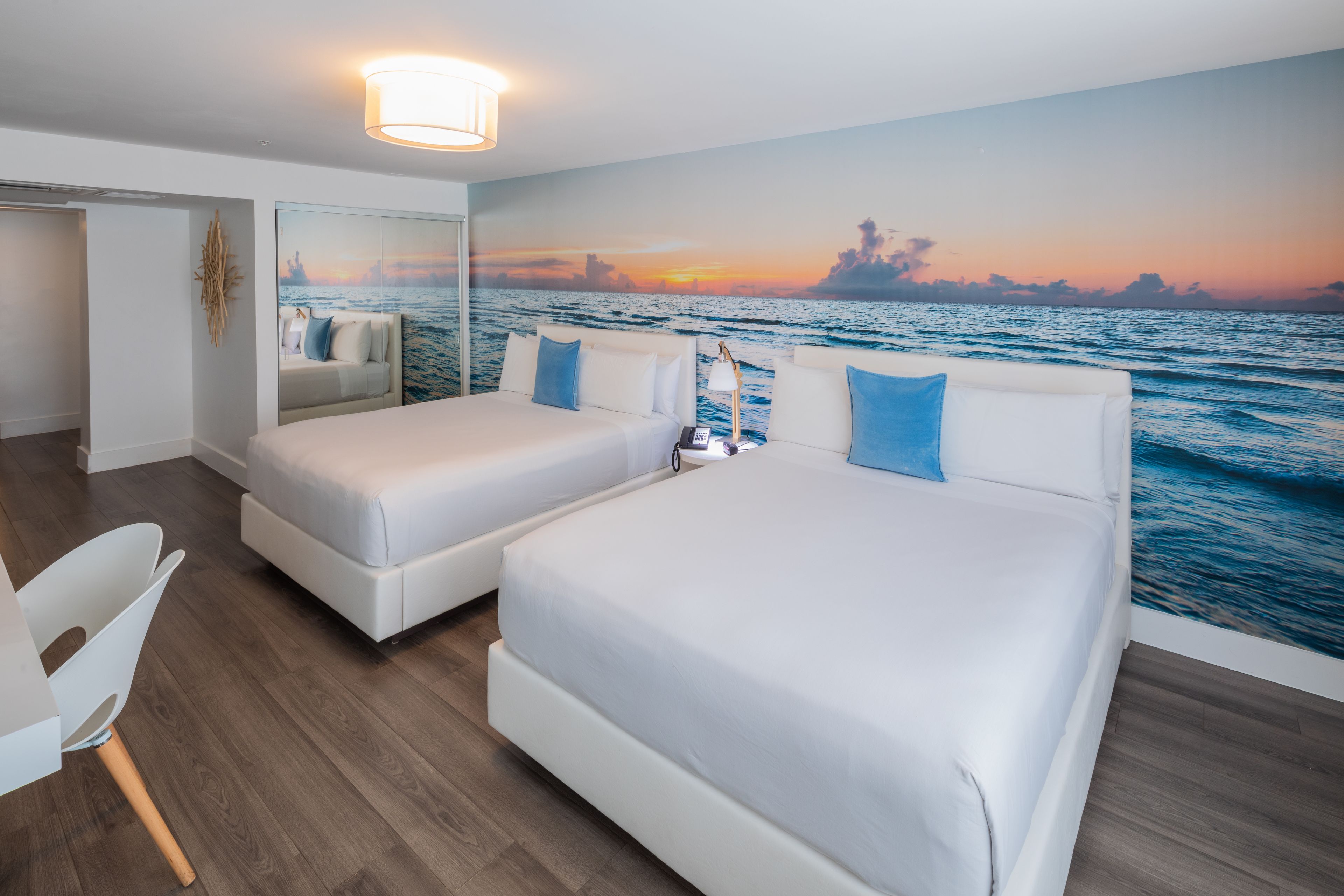 A hotel room with two beds, a white chair, a mirror, and a sunset mural on the wall.