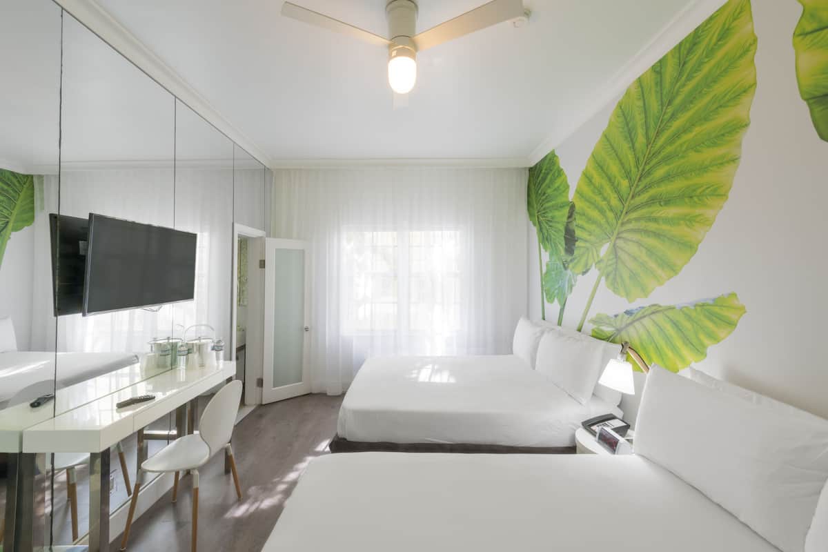 A hotel room with a bed, a desk with a TV, and large green leaves on the wall.