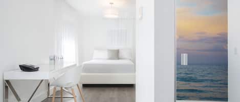 A modern hotel room with a white bed, a desk with a lamp, and a view of the sea through a large window.