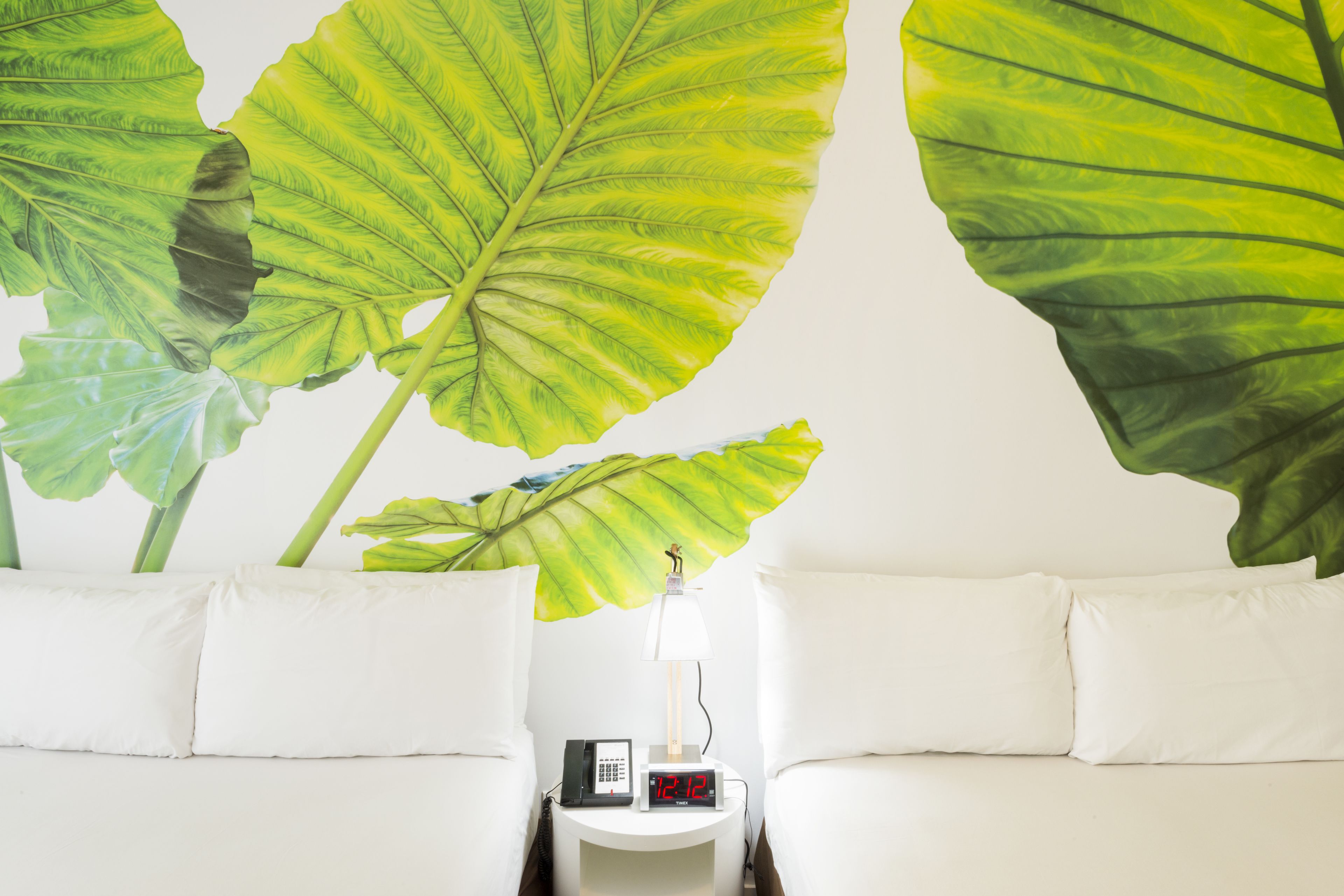 Une chambre d’hôtel avec un lit, des oreillers blancs, une table de chevet avec une lampe, une horloge et une fresque murale représentant de grandes feuilles vertes.