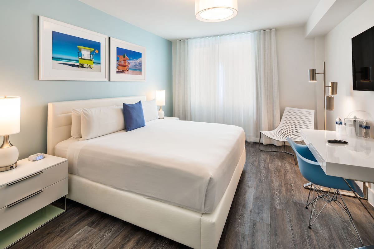 A hotel room with a bed, bedside table, lamp, desk, chair, and artwork on the wall.