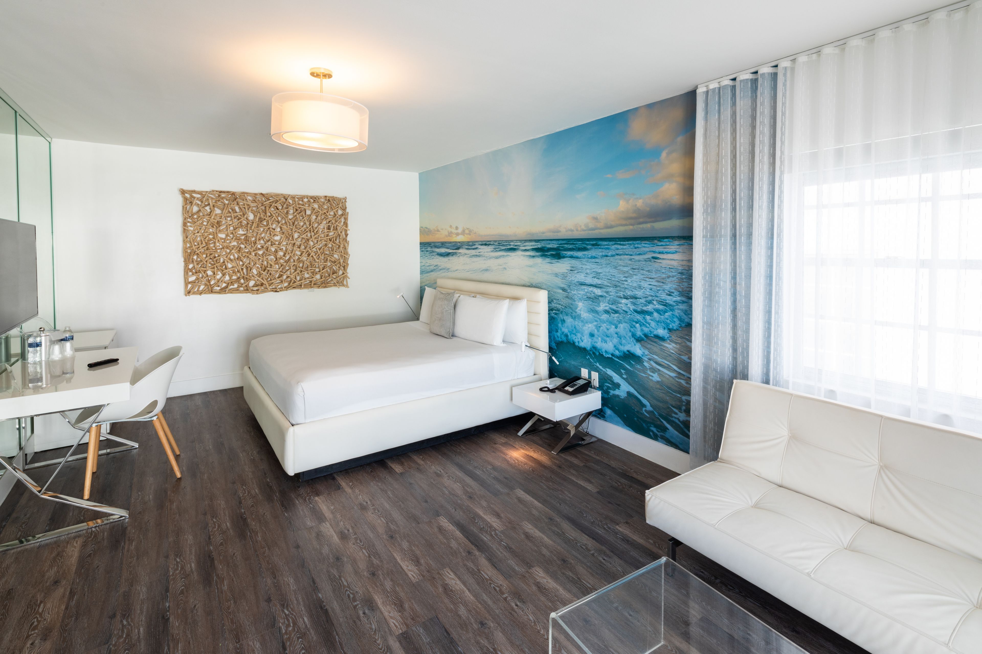 A modern hotel room with a large ocean mural, a white bed, a white sofa, a desk with a chair, and a glass coffee table.