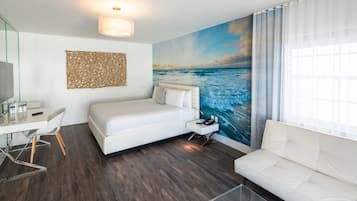 A modern hotel room with a large ocean mural, a white bed, a white sofa, a desk with a chair, and a glass coffee table.