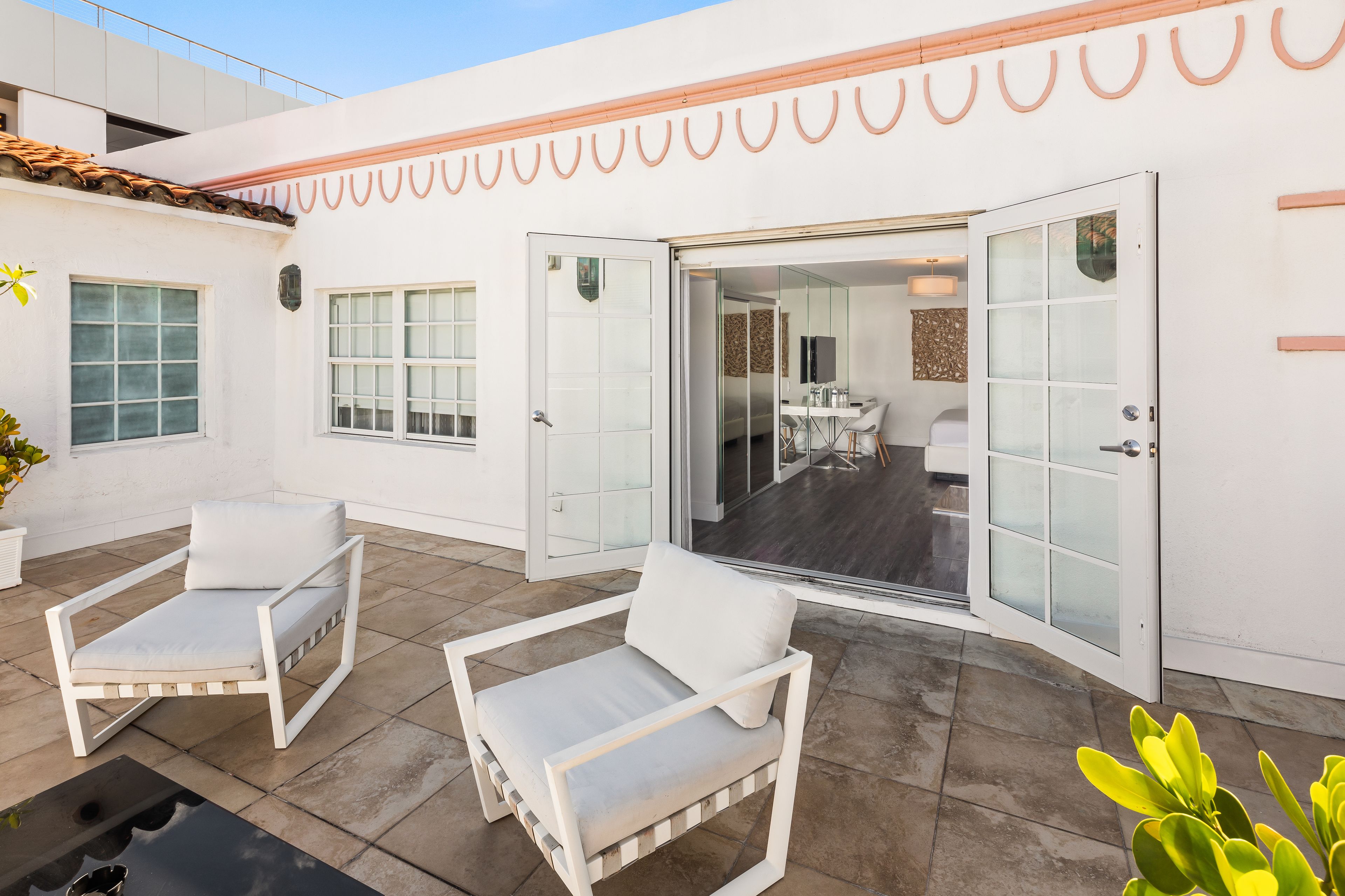 Un patio con dos sillas blancas acolchadas, un ventanal de vidrio que da a un comedor interior y una planta en maceta.