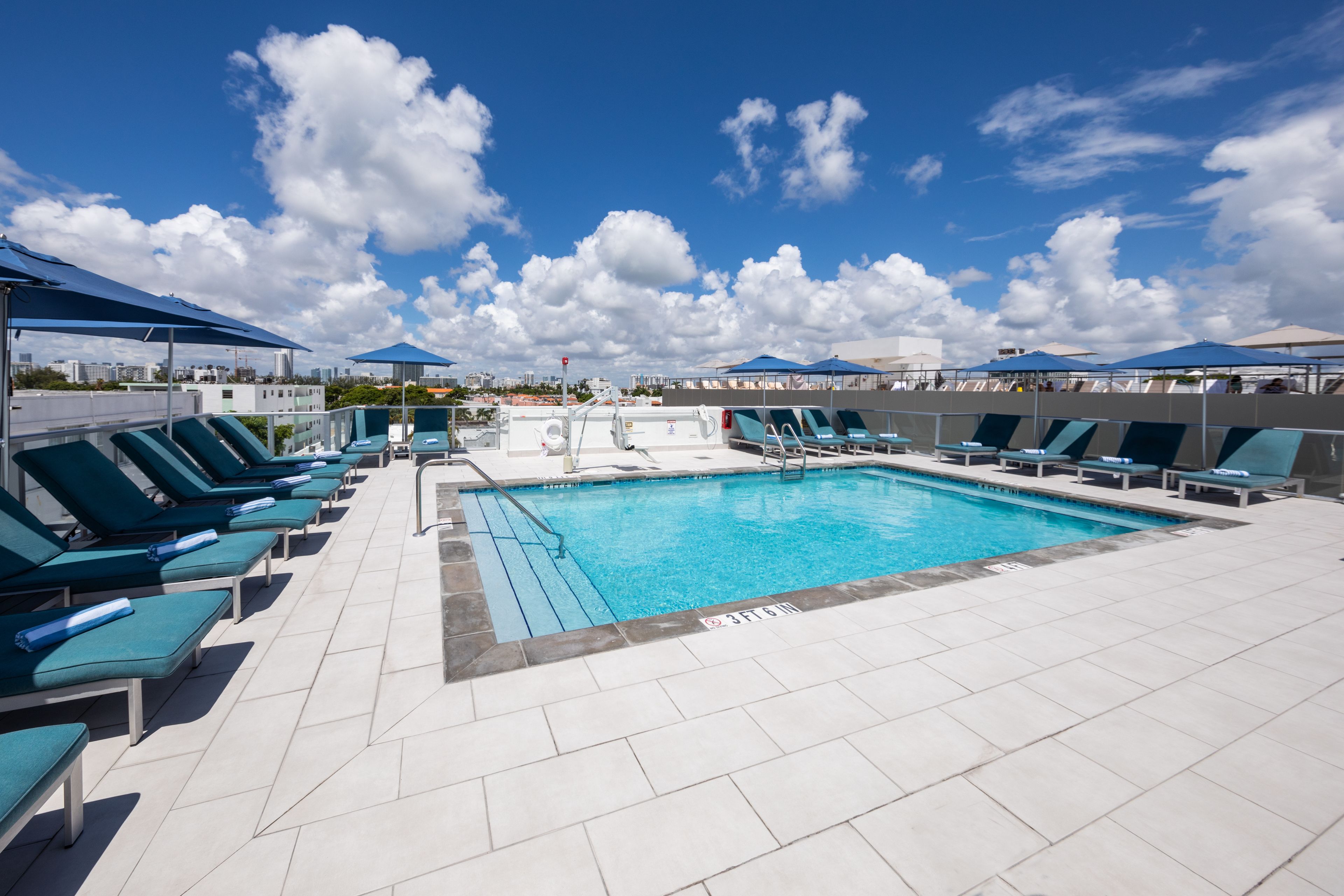 Un área de piscina con múltiples sillas de descanso y sombrillas, un cielo azul despejado y un piso de baldosas.