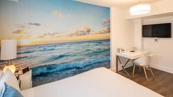 A bedroom with a large ocean mural, a bed, a desk with a chair, a TV, and a lamp.