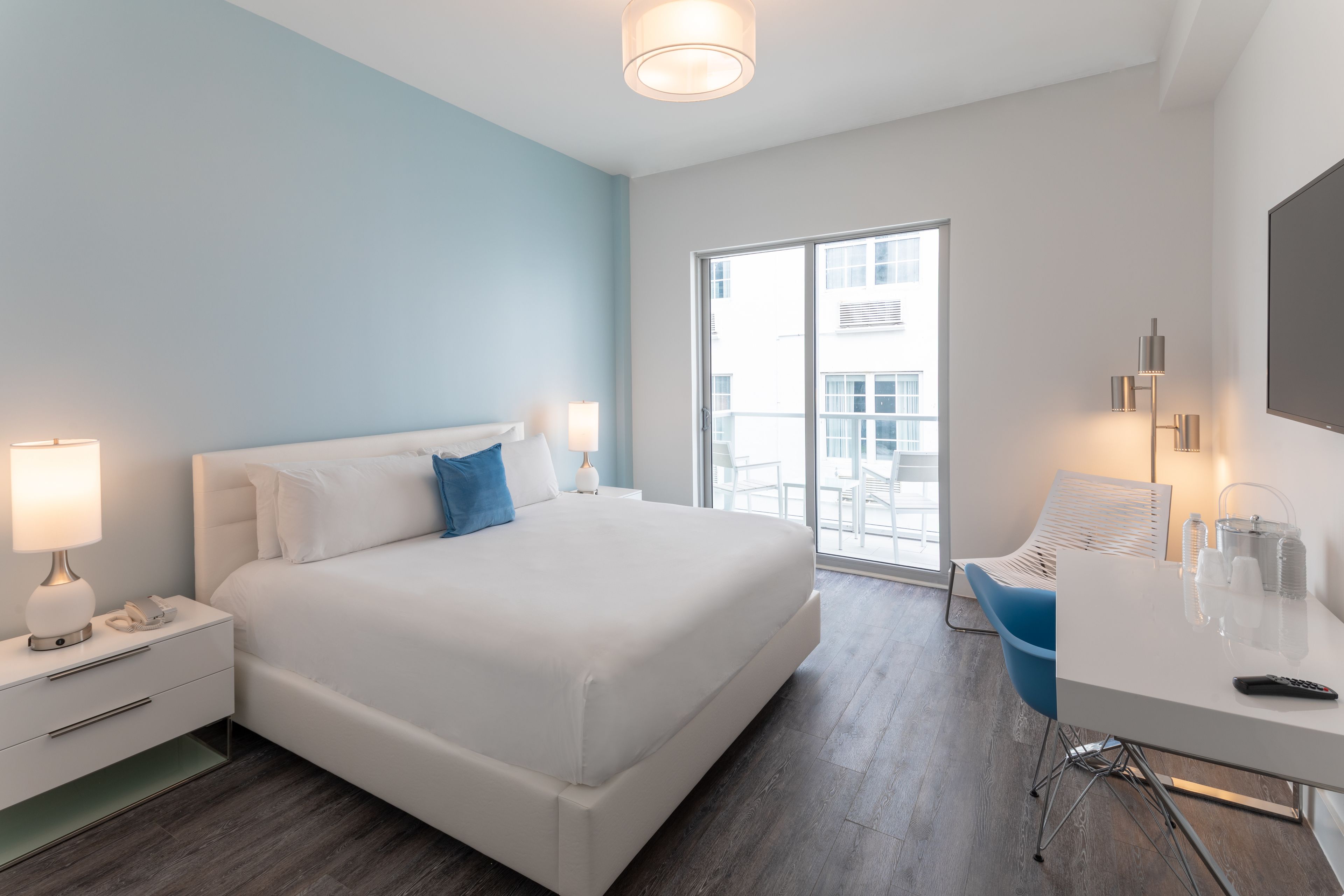 A modern hotel room with a large bed, a desk with a chair, a desk lamp, and a large window offering a view of the outside.