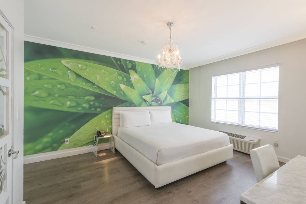 A bedroom with a large bed, a nightstand, a chandelier, a window, and a green leaf mural on the wall.