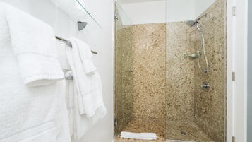 A glass-enclosed shower with a tiled wall, a glass shelf above the shower, and a white towel hanging on a rack.