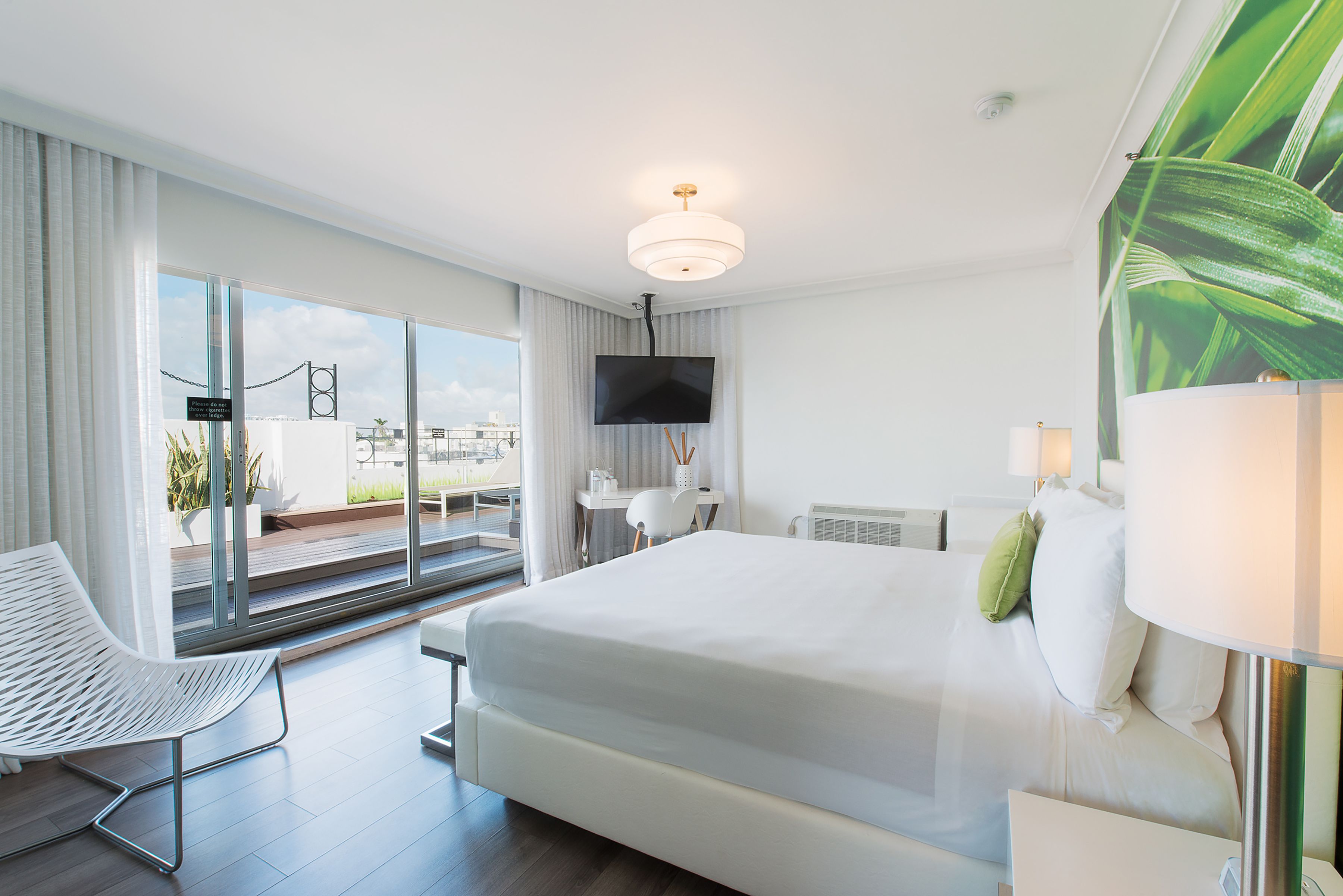 Una habitación de hotel moderna con una cama grande, un escritorio con televisor, una silla y un balcón con vistas a la ciudad.