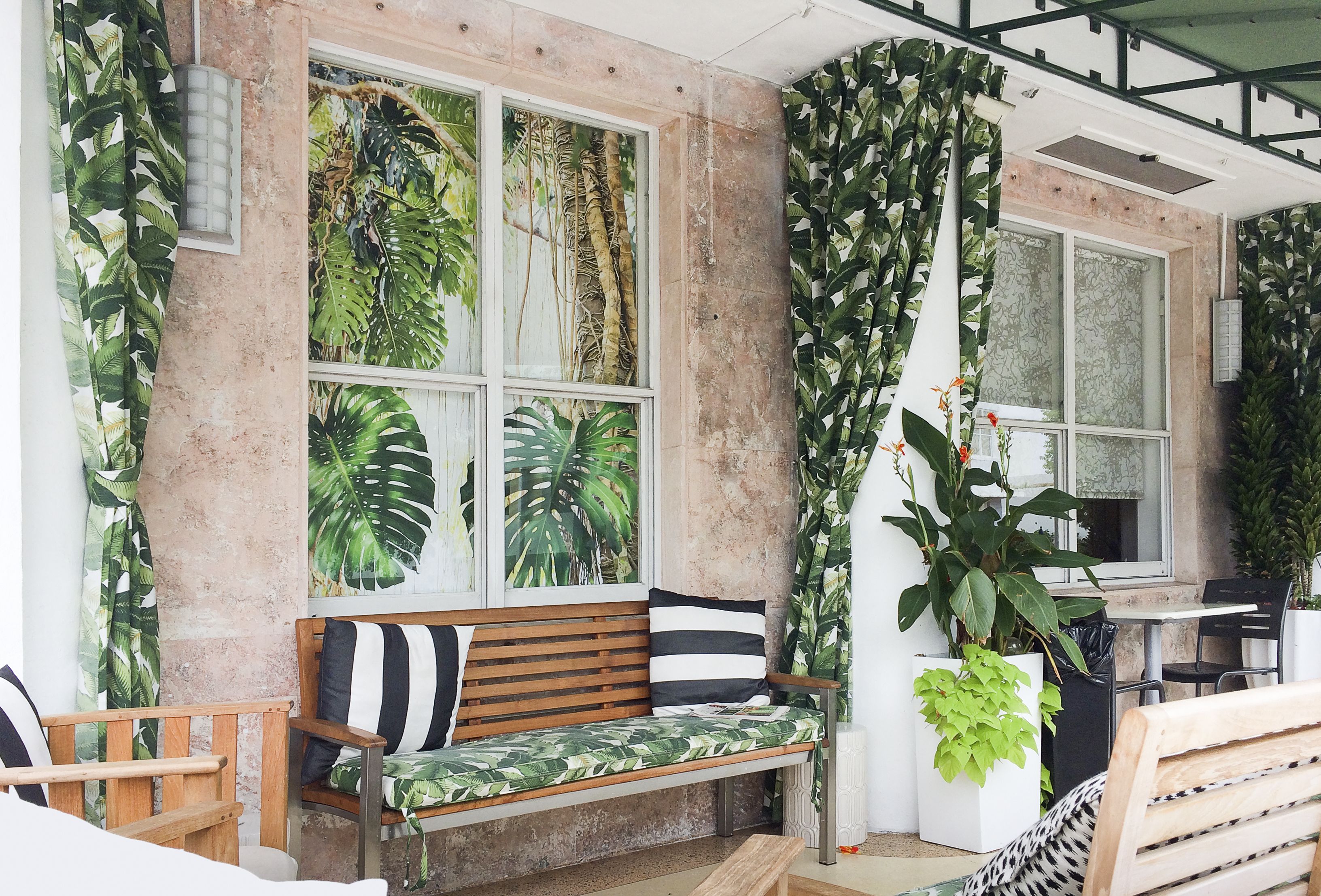 A room with a wooden bench, a table, and chairs. There are large windows with curtains, and a potted plant is visible.