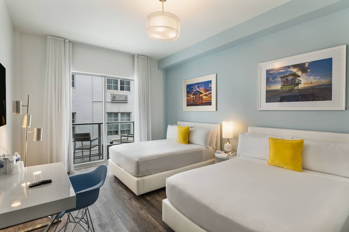 A hotel room with two beds, a desk, a chair, and a balcony with a view of the beach.