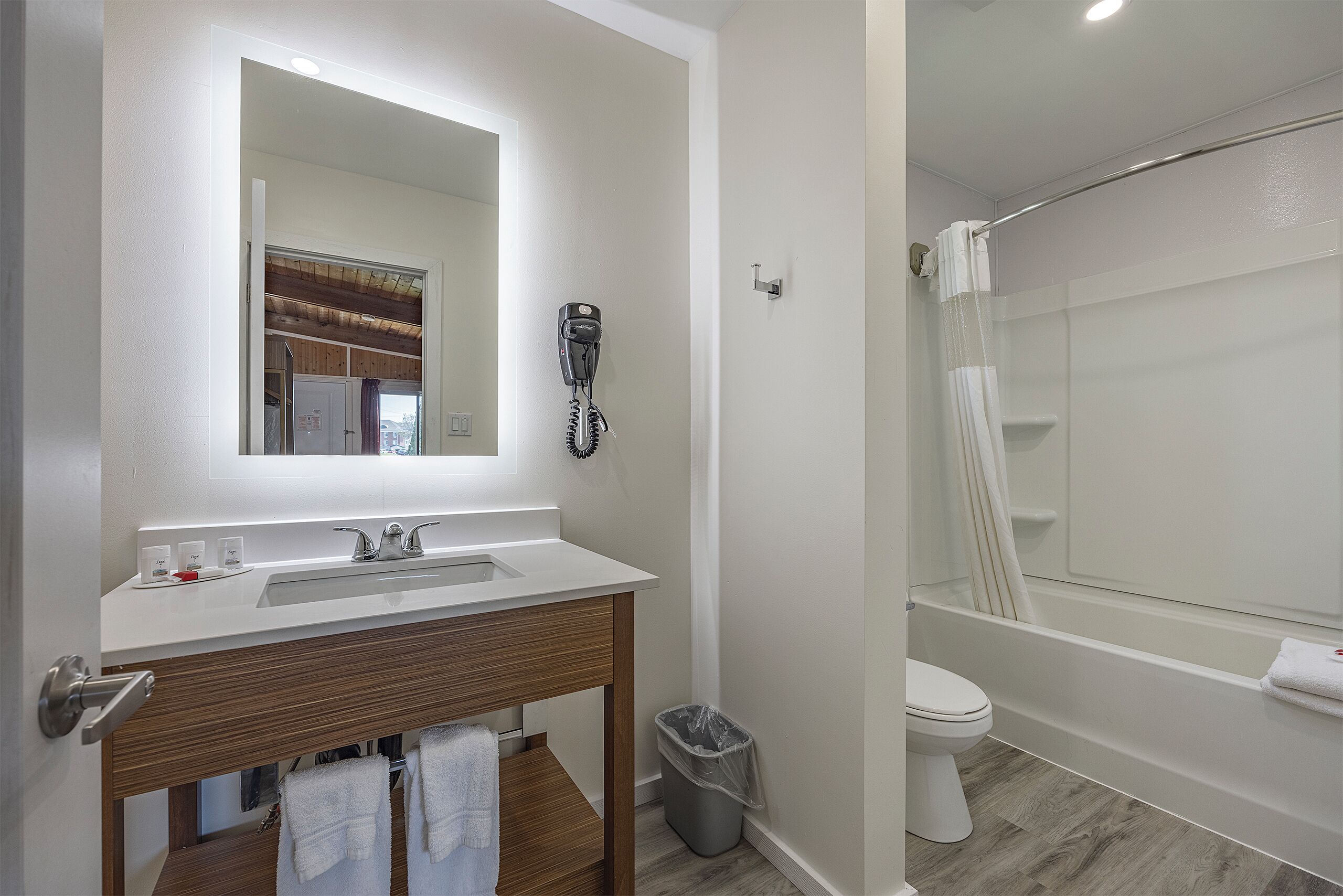 A bathroom with a white sink, wooden vanity, a hairdryer mounted on the wall, a toilet, and a bathtub with a shower curtain.