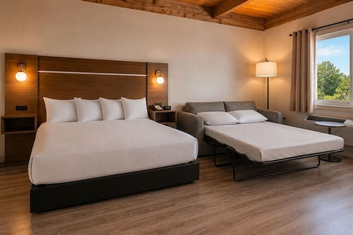 Una habitación de hotel con una cama grande, una cama más pequeña, un escritorio, una lámpara y una ventana con cortinas.