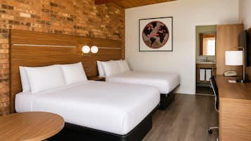 Quarto de hotel com duas camas, cabeceira de madeira, uma mesa redonda, uma escrivaninha com abajur, um mapa-múndi emoldurado e um banheiro visível através de uma porta aberta.