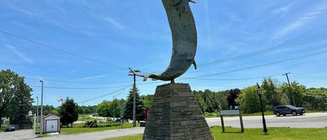 Una escultura de pez sobre un pedestal de piedra en un área con césped, con un camino y árboles al fondo.