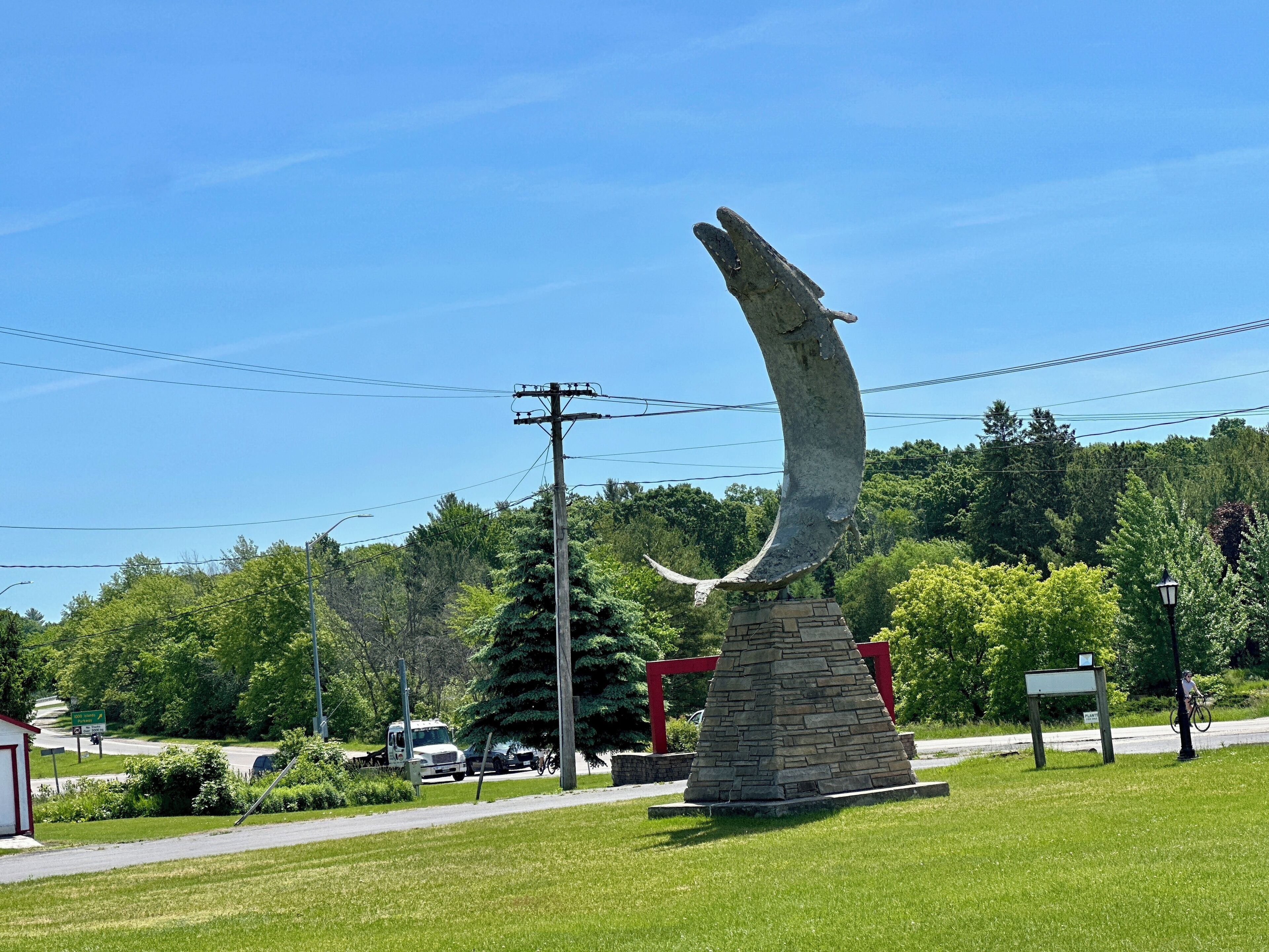 A sculpture of a crescent moon on a stone pedestal in a grassy area with a road and trees in the background.