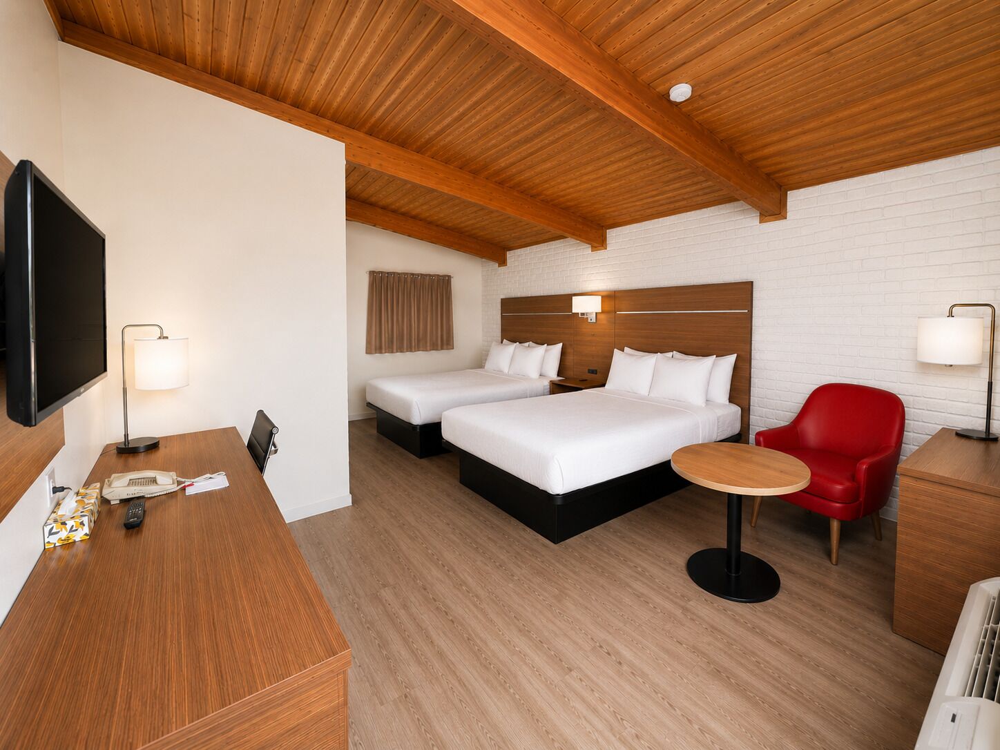 Une chambre d’hôtel avec un plafond en bois, deux lits, une télévision, un bureau avec une lampe, une chaise rouge et une table ronde.
