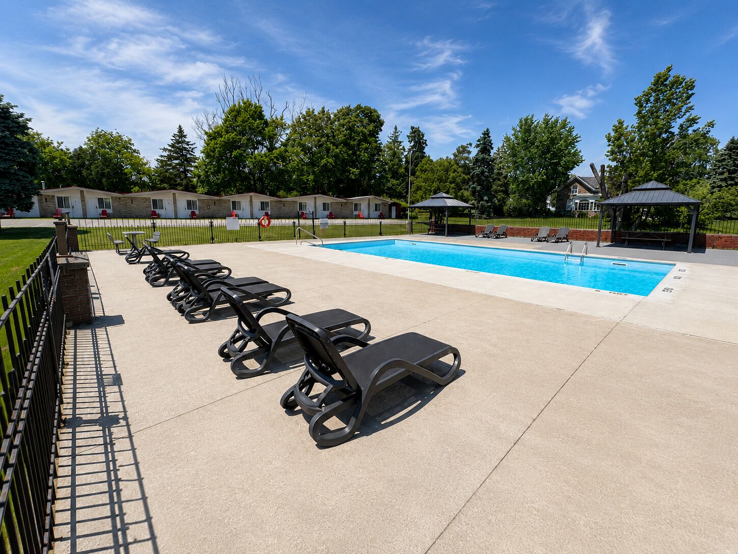 Una piscina con sillones de descanso, un pabellón y un área cercada con un edificio al fondo.