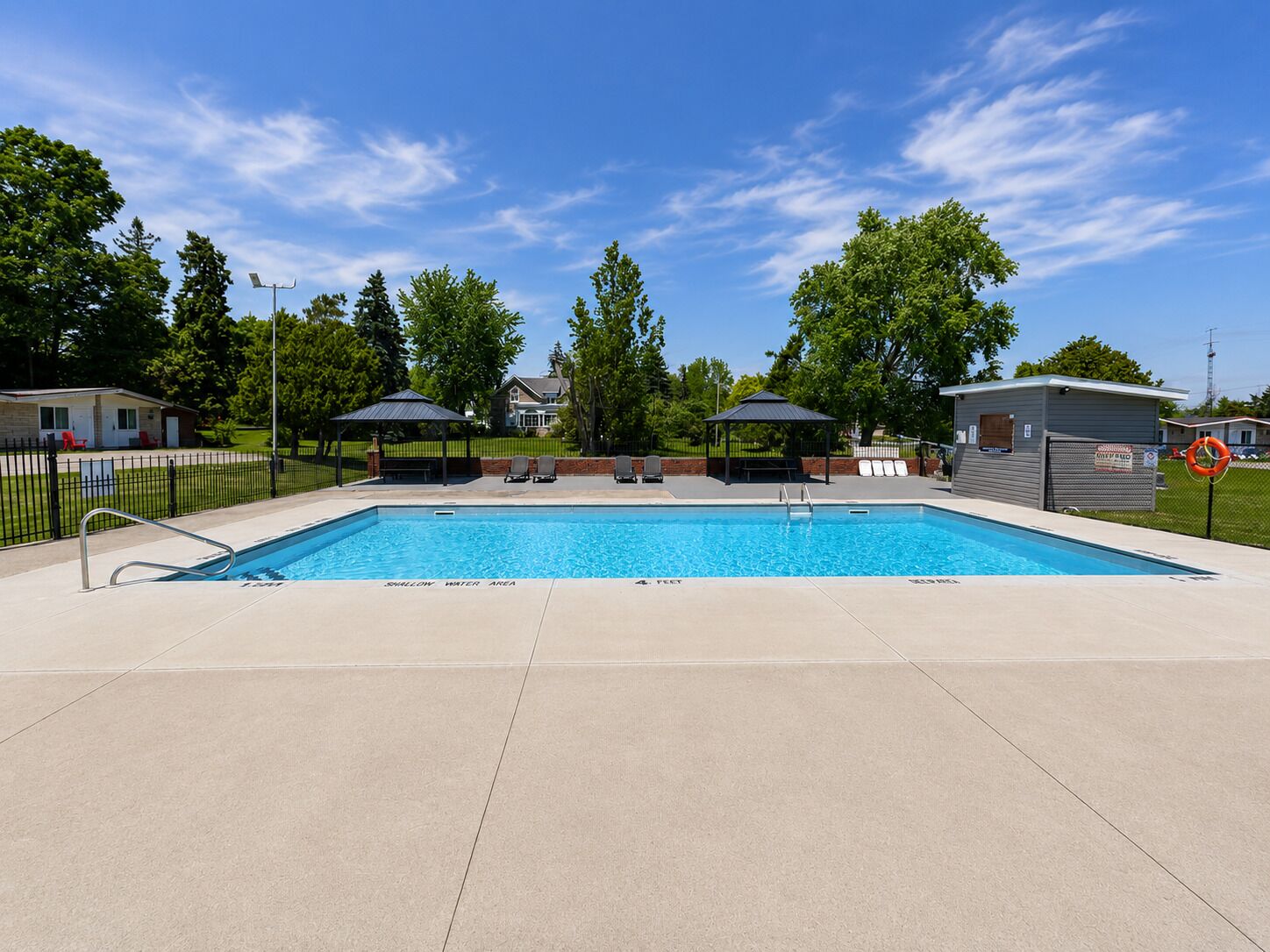 A rectangular swimming pool with a shallow water area, surrounded by a paved deck, two covered seating areas, and a fenced area with lounge chairs.