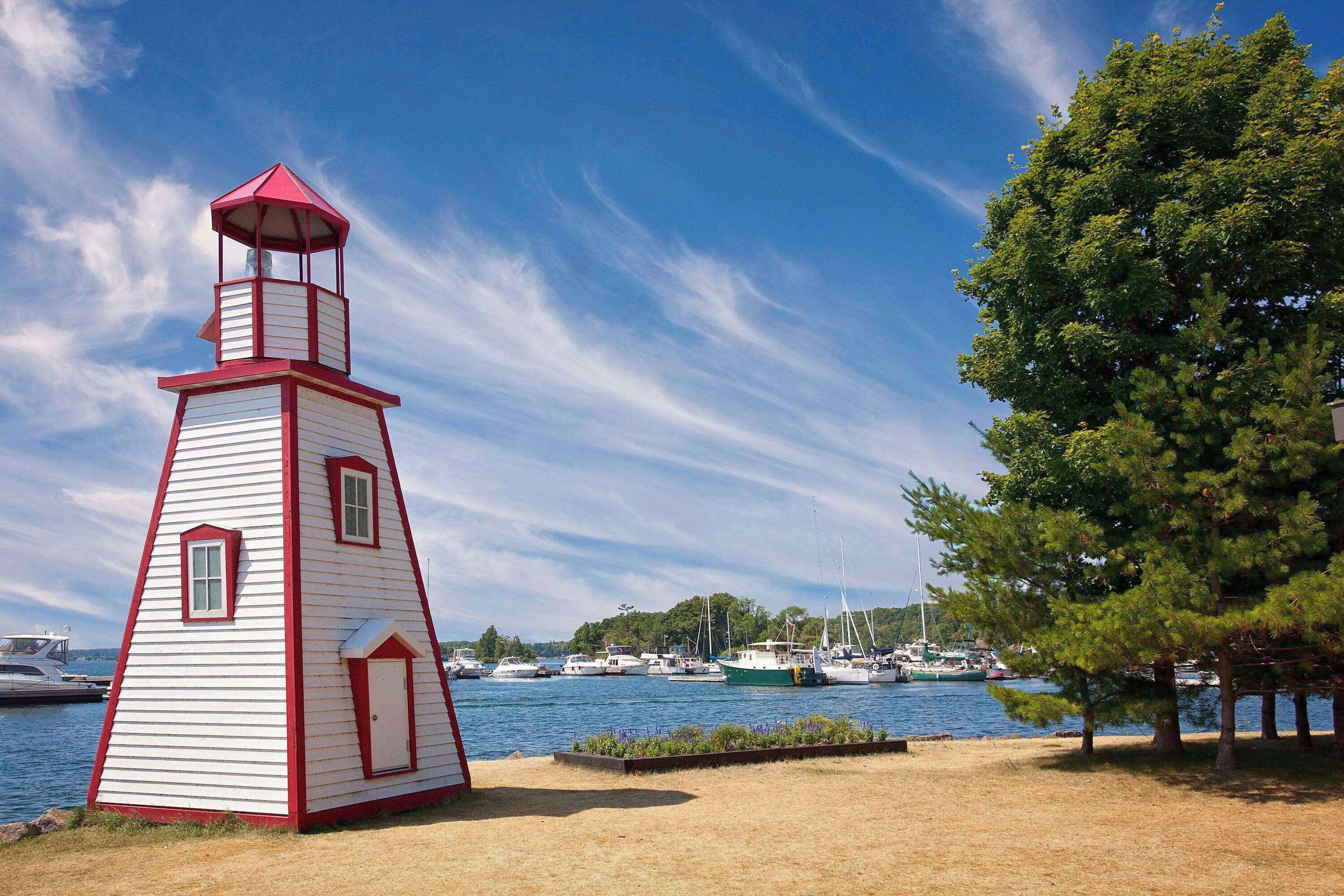 Pequeña estructura de faro blanco y rojo cerca del agua, con barcos y árboles.
