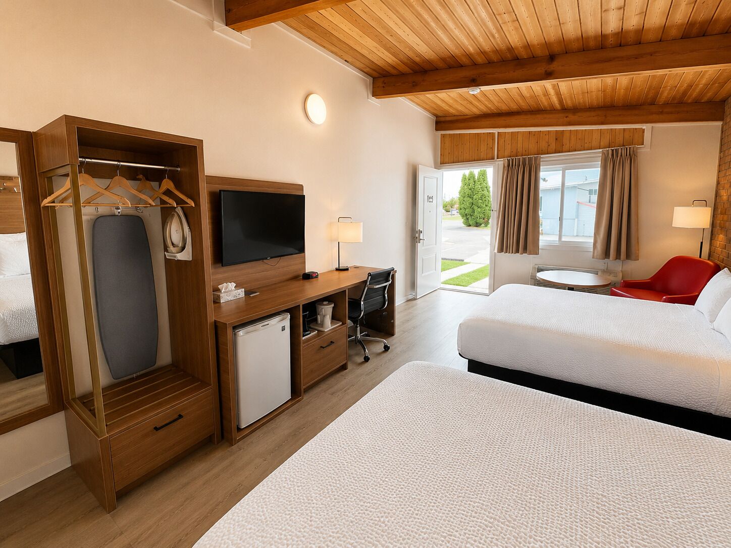 A hotel room with a bed, a desk with a TV, a mini-fridge, a coat rack, and a red chair.