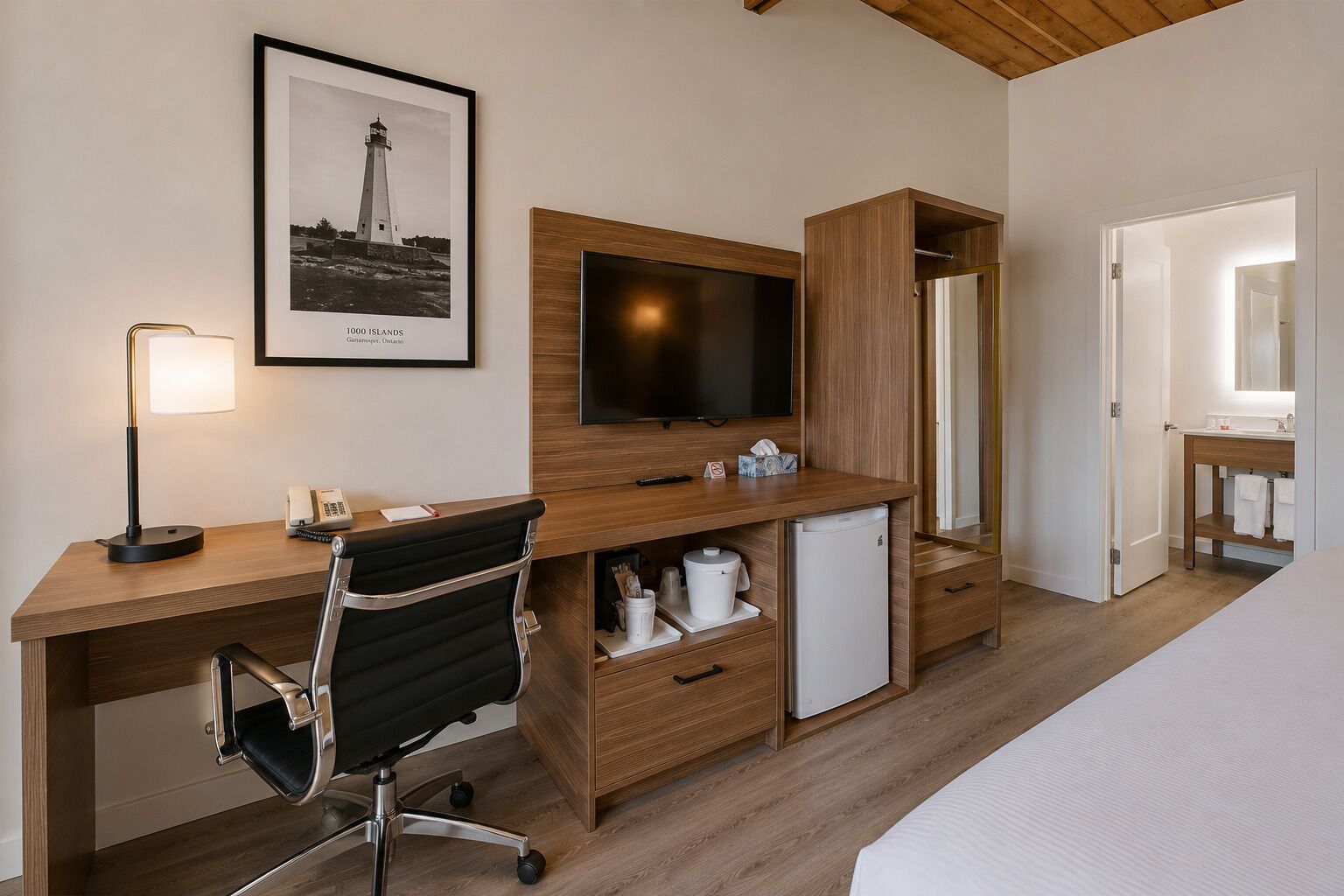 Una habitación de hotel con un escritorio de madera, una silla de oficina negra, un televisor, un refrigerador pequeño, una mesita de noche y una cama.