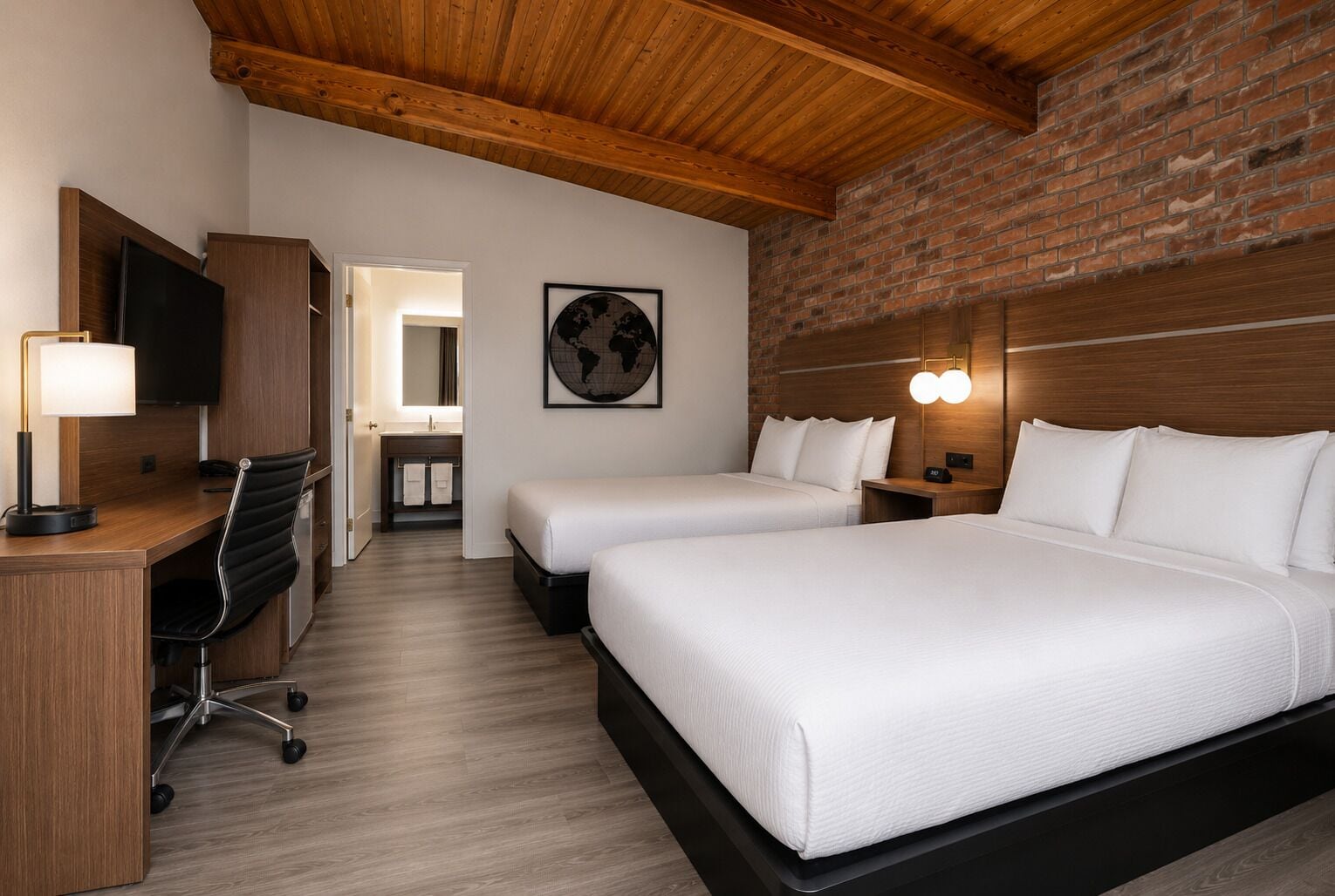 A hotel room with a wooden ceiling, a brick wall, a desk with a lamp, a TV, two beds, a bathroom, and a world map on the wall.