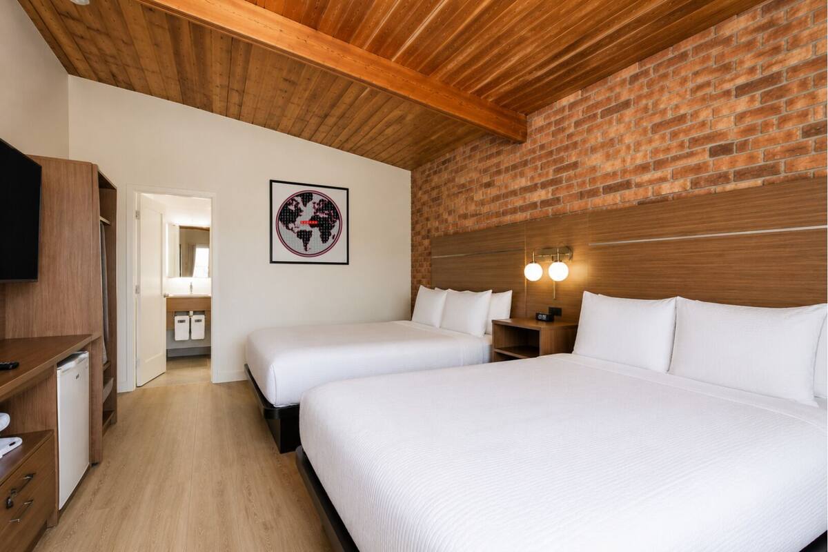 A hotel room with two beds, a wooden ceiling, a brick wall, and a framed picture on the wall.