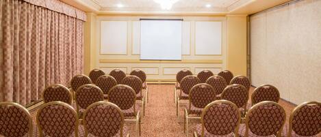 Un salón de conferencias con filas de sillas, una pantalla de proyector y piso alfombrado.