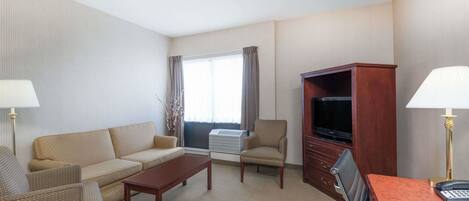 A hotel room with a sofa, armchair, coffee table, TV stand, and desk with a lamp.