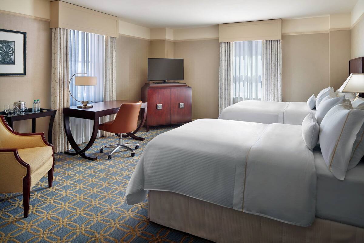 A hotel room with two beds, a desk with a TV, a chair, a wardrobe, and a patterned carpet.