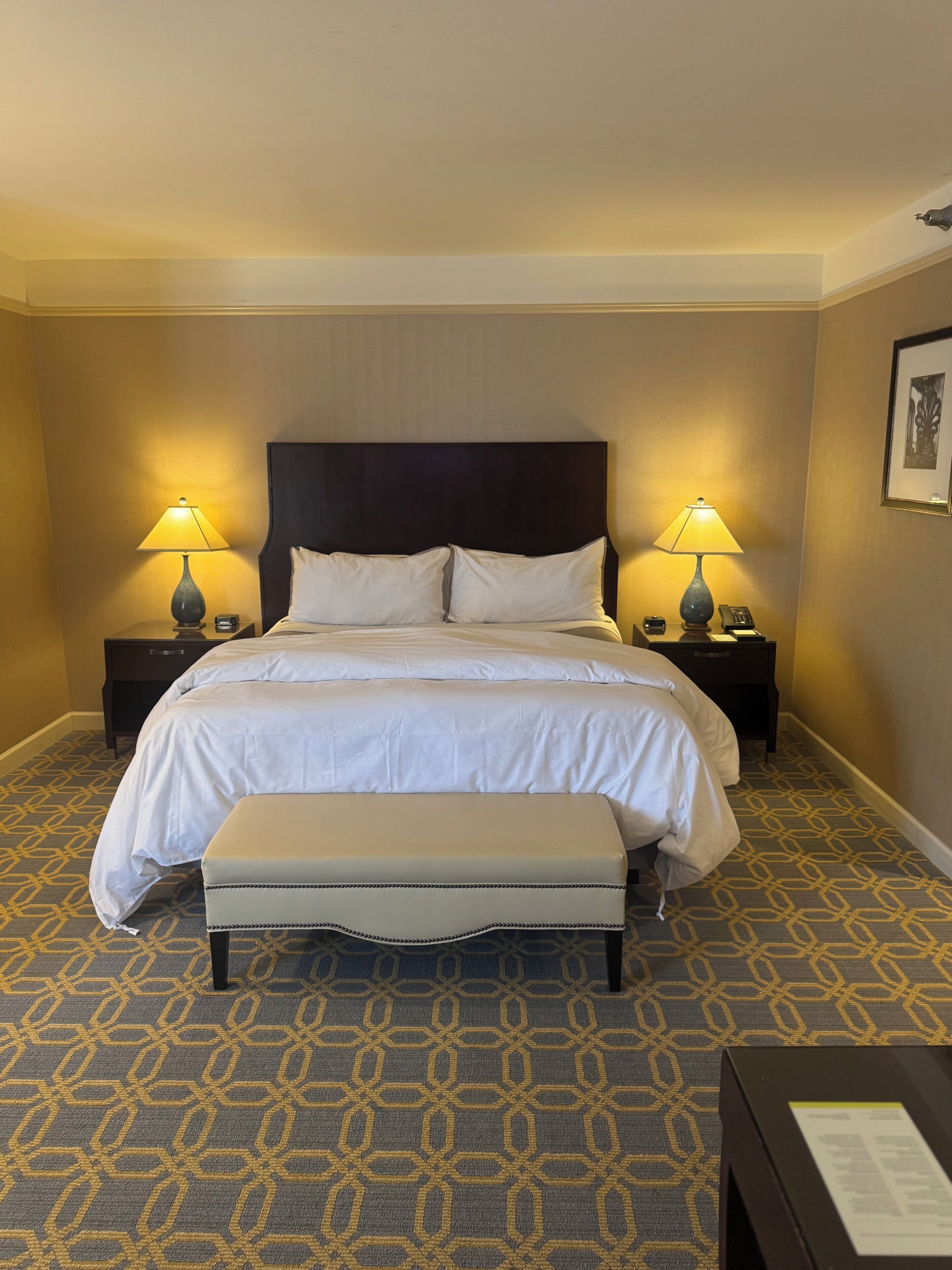 A hotel room with a large bed, two bedside tables with lamps, a bench, patterned carpet, and a framed picture on the wall.