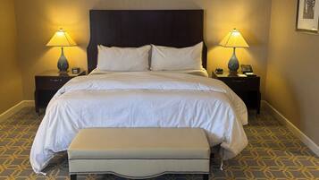 A hotel room with a large bed, two bedside tables with lamps, a bench, patterned carpet, and a framed picture on the wall.