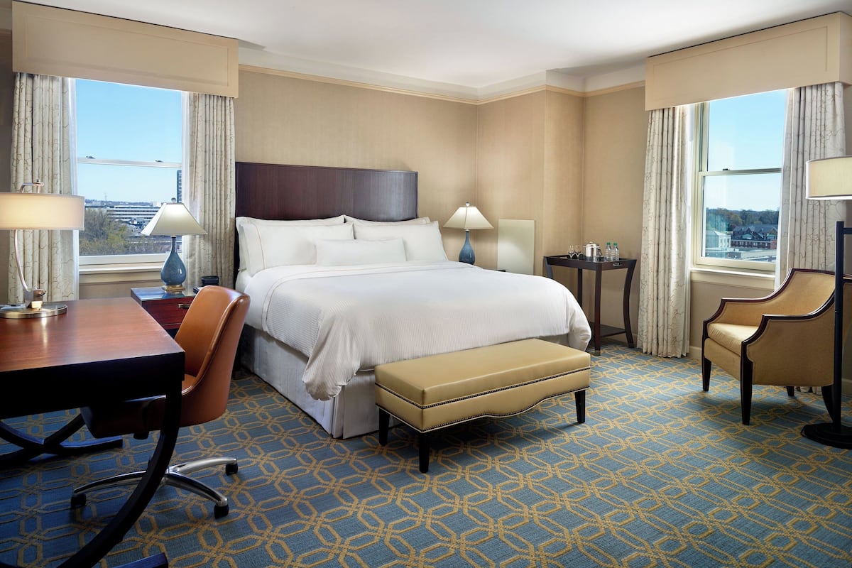 A hotel room with a large bed, a desk with a chair, a small table with a lamp, and a view of the city through the windows.