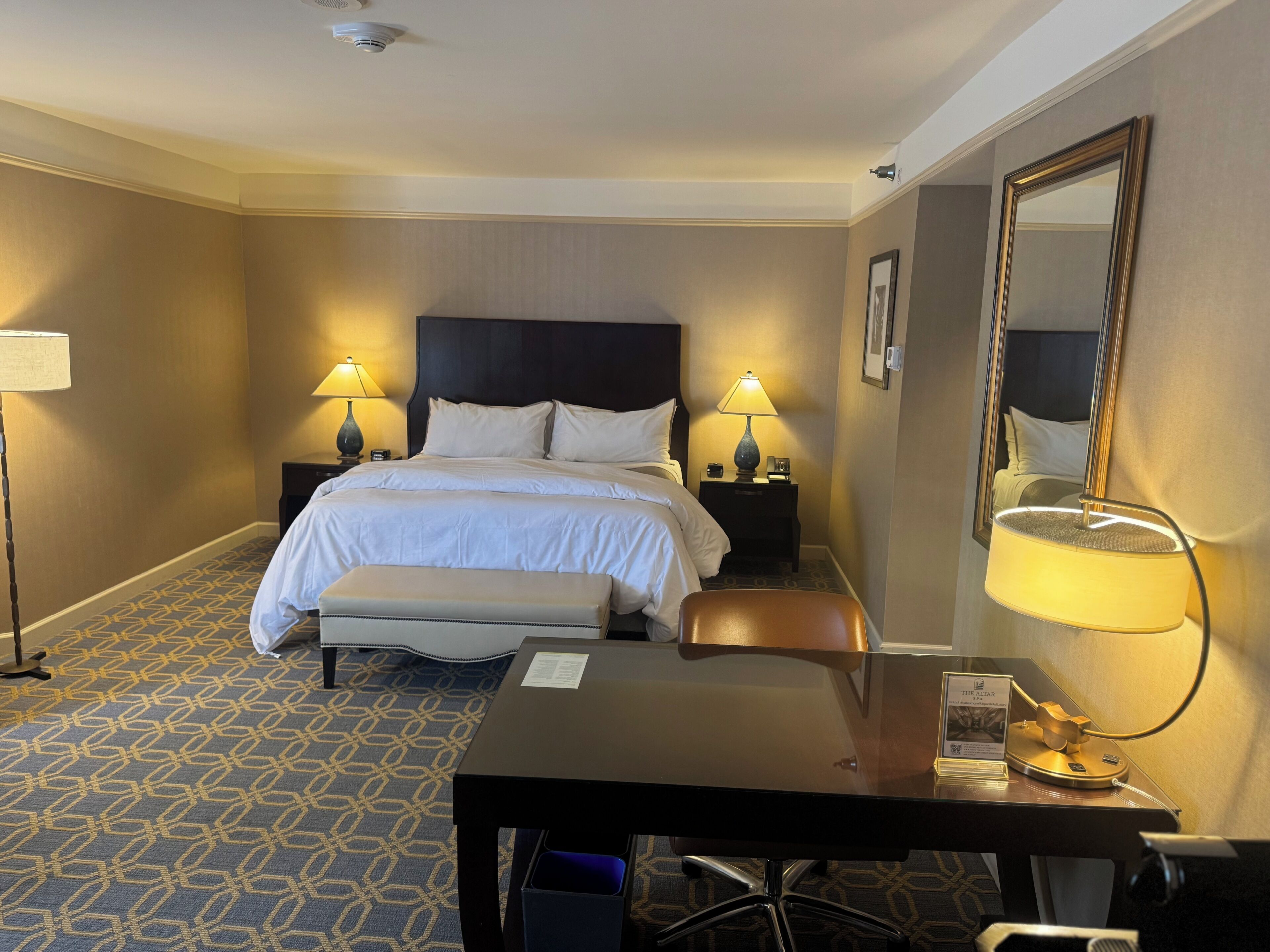A hotel room with a bed, bedside lamps, a desk, and a mirror.