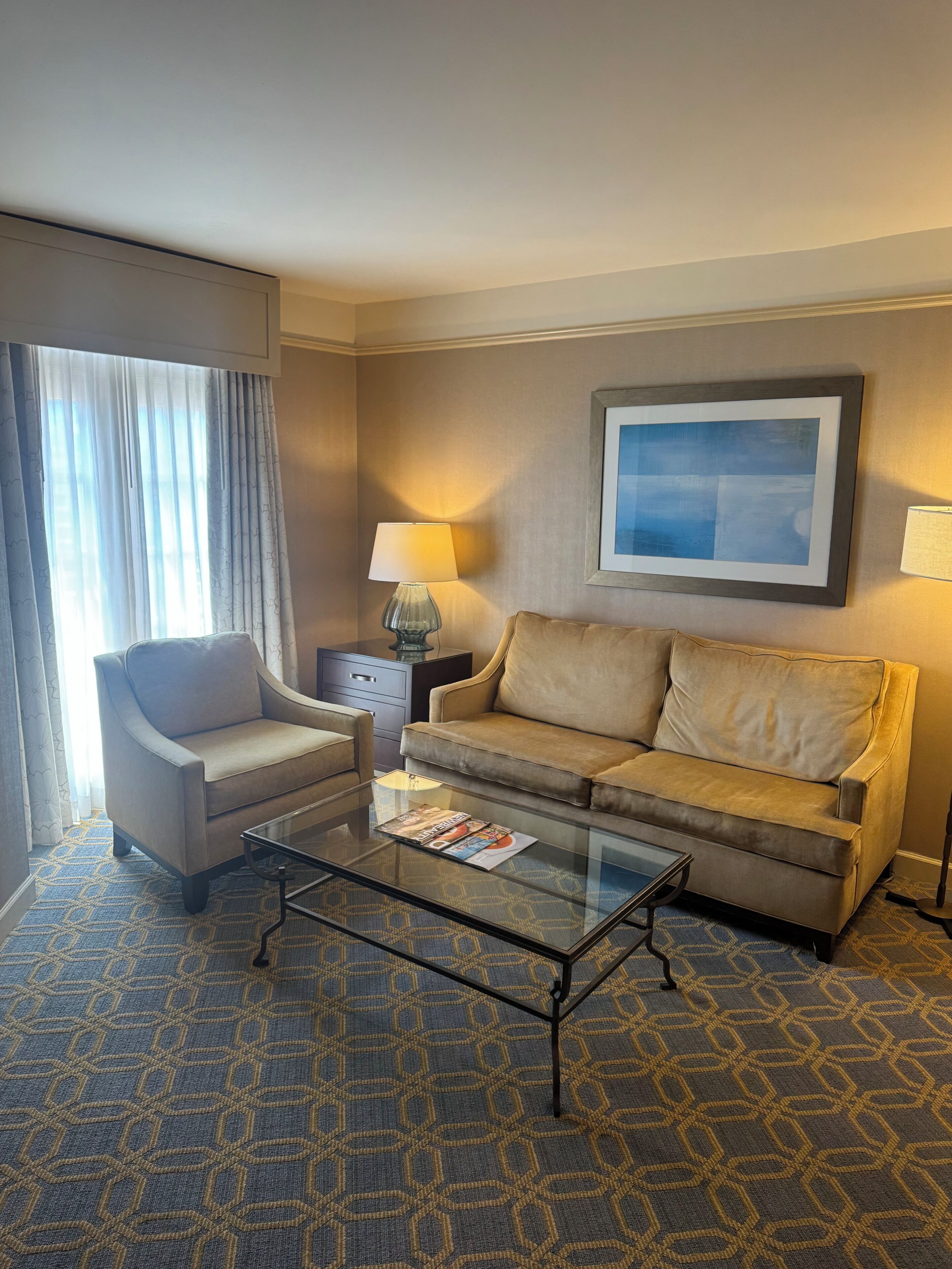 A hotel room with a sofa, armchair, glass coffee table, and a lamp.