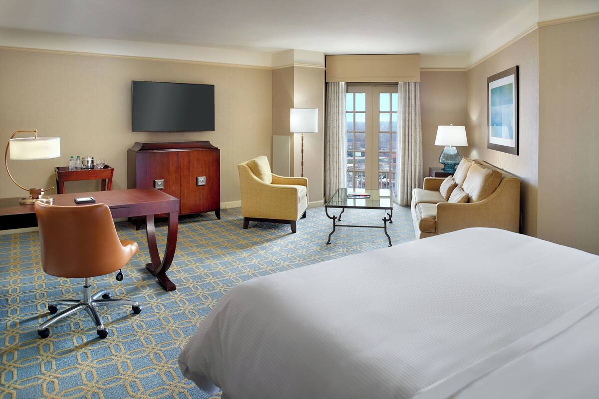 A hotel room with a large bed, two armchairs, a desk with a flat-screen TV, a dresser, and a view of the city through a large window.