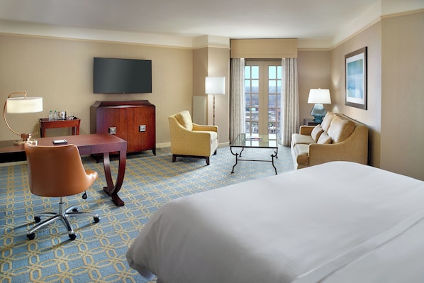 A hotel room with a large bed, two armchairs, a desk with a flat-screen TV, a dresser, and a view of the city through a large window.