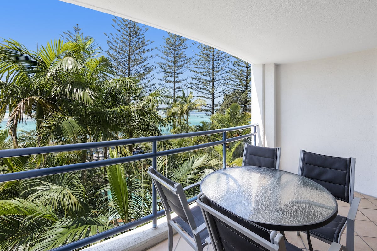Un balcon doté d’une table et de chaises en verre, offrant une vue sur un paysage tropical avec des palmiers et sur l’océan.