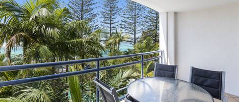 Un balcón con una mesa y sillas de vidrio, con vistas a un paisaje tropical con palmeras y al océano.