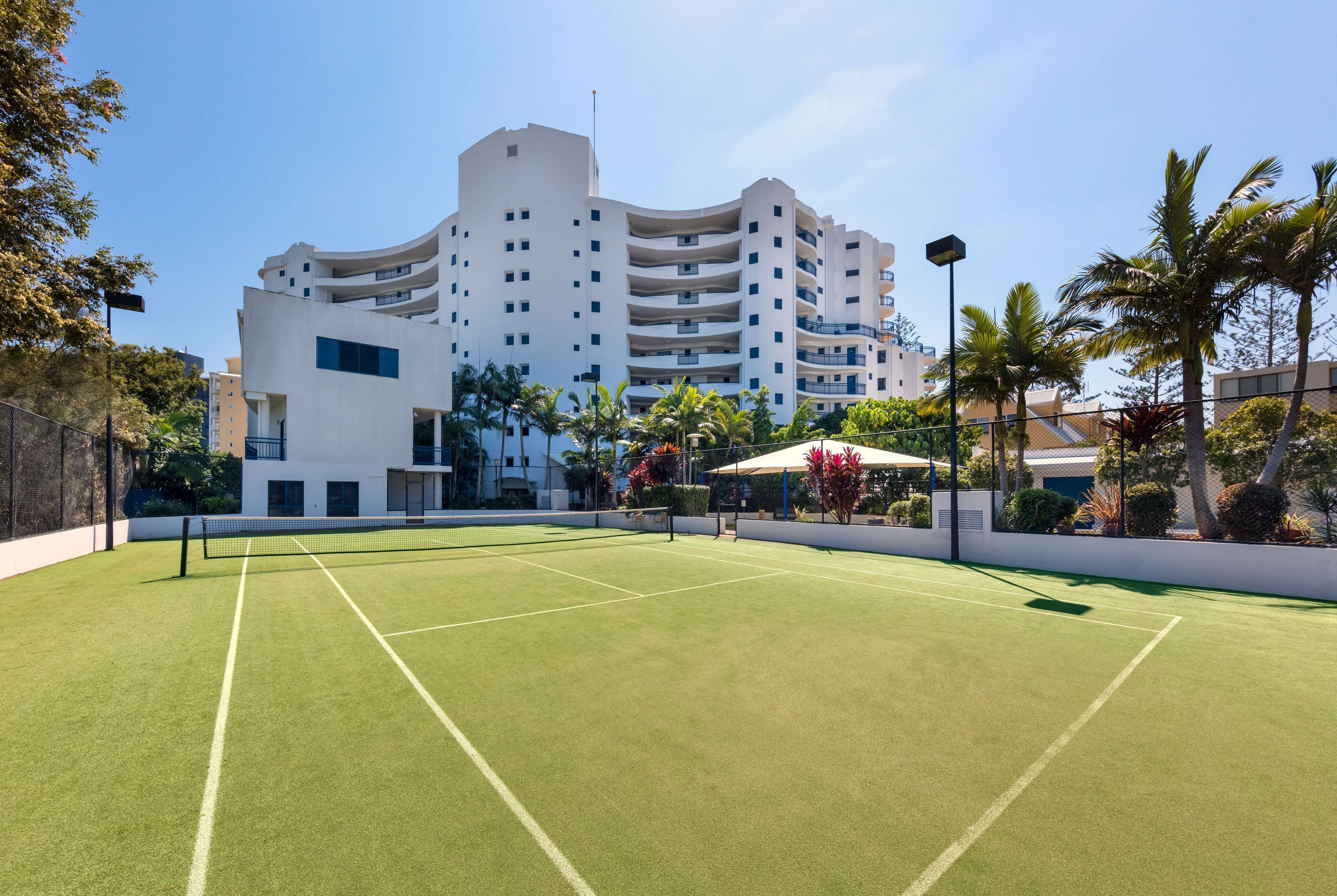 Een tennisbaan met een net, omringd door een hek, en een gebouw met meerdere balkons op de achtergrond.